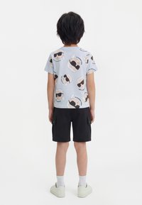 Copil stând cu fața spre spate, purtând un tricou bleu deschis cu fețe de desene animate repetate, purtând ochelari de soare, și pantaloni scurți cargo negri.