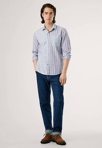 Camisa de botões com riscas azul claro e branco, com um bolso no peito, combinada com jeans azul escuro e sapatos castanhos.