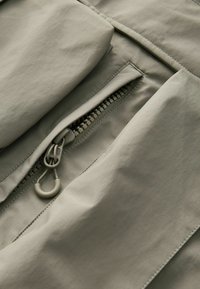 Outerwear de culoare gri deschis, cu un buzunar cu fermoar, textură netedă și un model subtil în grilaj pe material. Include hardware pentru cursa ajustabilă a fermoarului.