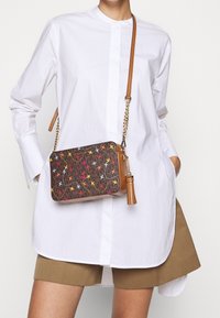 Borsa a tracolla in tessuto marrone con uccelli ricamati colorati, tracolla in catena dorata e dettaglio a nappina, abbinata a una camicia bianca e pantaloni corti khaki.