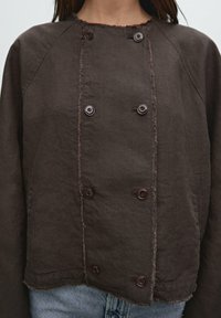 Brun knapad linjacka med råa kanter, bärs över ljusblå denimjeans, visad från bröstet till höfterna.