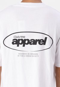 Camiseta blanca de algodón con un gran logo ovalado negro que incluye el texto "apparel" y "Club1990". Debajo, en un sutil texto gris, se lee: "DISEÑADO EN DINAMARCA POR LA SOCIEDAD JJ DENIM."