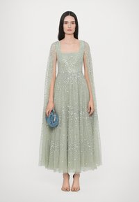 Lange, lichtgroene jurk met paillettenversieringen en een doorzichtige cape. Gecombineerd met een blauwe, gestructureerde tas en nude schoenen.