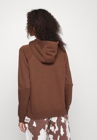 Brown hoodie in soft material with a drawstring hood. Features raglan sleeves and a straight hem. Worn with patterned, light brown and white pants.
