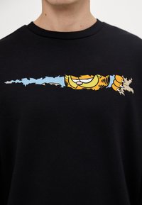 Felpa nera con una grafica cartoon colorata di un gatto che sbircia attraverso una sezione strappata, con uno sfondo blu e espressioni facciali dettagliate.