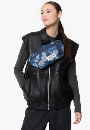 Gilet en cuir noir avec une fermeture éclair à l'avant, porté sur un pull sombre. Une ceinture banane bleu et blanc tie-dye est portée en bandoulière, comportant une fermeture éclair et un logo.