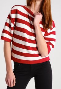 Rot-weiß gestreifter Strickpullover mit weit ausgeschnittenem Halsausschnitt und kurzen Ärmeln, kombiniert mit schwarzen Hosen. Der Stoff hat eine strukturierte Oberfläche.