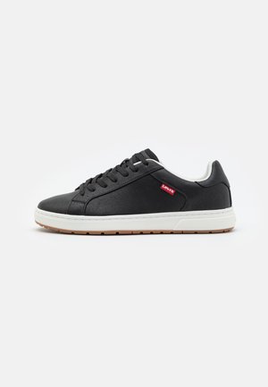 Zapatilla negra de cuero con una entresuela blanca, superficie texturizada, puntera redonda y logo rojo en el lateral. La suela de goma proporciona tracción.