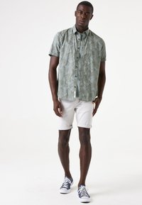 Korte mouwen shirt in lichtgroen met een bladmotief, gecombineerd met witte shorts met omgeslagen rand en blauwe sneakers. Beide items hebben een casual ontwerp.