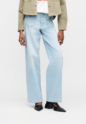 Φαρδιά τζιν παντελόνια σε ανοιχτό γαλάζιο χρώμα, φορεμένα με μαύρα μυτερά παπούτσια με χαμηλό τακούνι, μπεζ cropped σακάκι και λευκό μπλουζάκι με γραφικό σχέδιο.