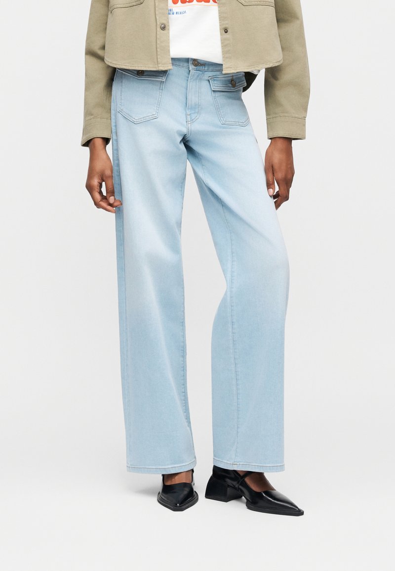 Φαρδιά τζιν παντελόνια σε ανοιχτό γαλάζιο χρώμα, φορεμένα με μαύρα μυτερά παπούτσια με χαμηλό τακούνι, μπεζ cropped σακάκι και λευκό μπλουζάκι με γραφικό σχέδιο.