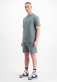 Lichtgrijs-groene t-shirt met korte mouwen, gecombineerd met bijpassende shorts. Bijzonder logo-opdruk op de mouw van het shirt en de zoom van de shorts. G gedragen met een paar gemusterde sneakers.