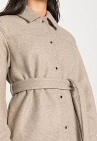Cappotto di lana beige con un ampio colletto, bottoni a scatto neri e una cintura coordinata che stringe in vita, indossato da una persona con capelli scuri.