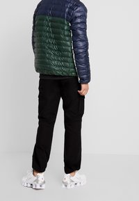 Fodrad jacka i mörkgrön och marinblå med kviltad textur. Svarta cargo-byxor och vita sportskor, vy bakifrån.