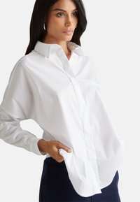 Camicia bianca a maniche lunghe con colletto classico, tasca frontale discreta e vestibilità rilassata, abbinata a pantaloni scuri. Tessuto morbido, design minimalista.