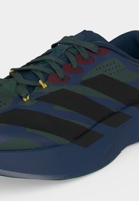 Κοντινή λήψη ενός αθλητικού παπουτσιού σε σκούρο μπλε και πράσινο με μαύρες ρίγες, κόκκινες λεπτομέρειες κοντά στα κορδόνια και κίτρινες άκρες κορδονιών.