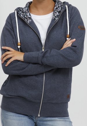 Personne portant un sweat à capuche gris foncé ouvert avec cordons blancs et doublure de capuche à motif, bras croisés sur un t-shirt blanc et un jean clair.