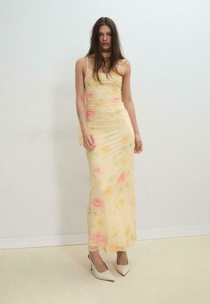 Junge Frau mit langen dunklen Haaren, die ein gelbes, geblümtes Trägerkleid und weiße High Heels trägt, steht vor einer schlichten Wand.