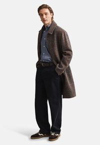 Brūns pleds virsjaka, zilas pogājamas krekls, tumšas bikses un brūnas sporta kurpes. Virsjakai ir izgriezta apkakle un fakturēts audums.