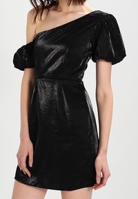 Kvinna som bär en glänsande svart enaxlad miniklänning med puffade korta ärmar, stående mot en enkel ljus bakgrund.