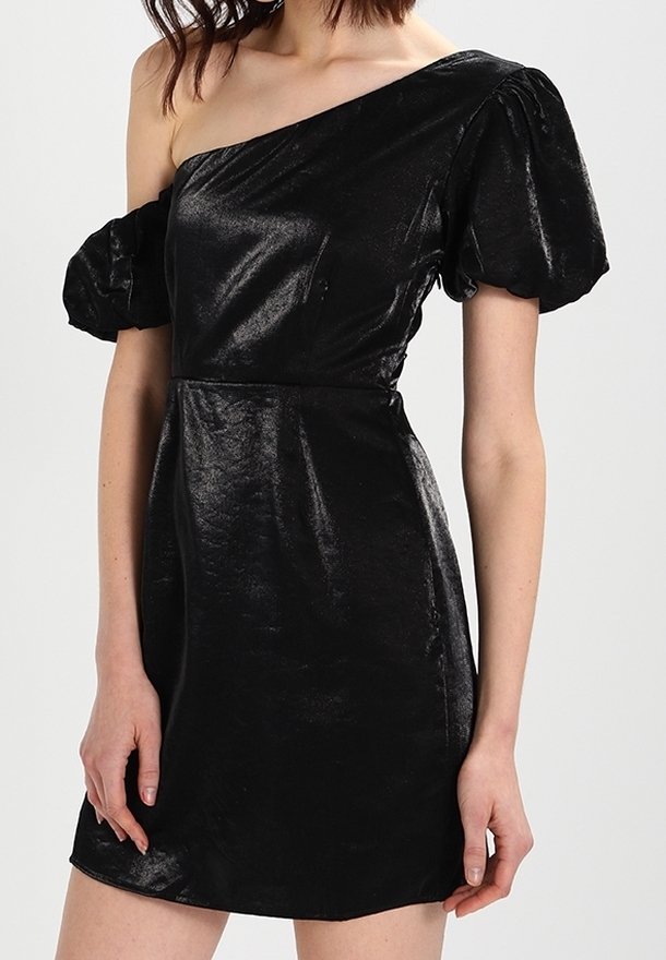 Kvinna som bär en glänsande svart enaxlad miniklänning med puffade korta ärmar, stående mot en enkel ljus bakgrund.