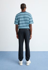 Ein Mann mit geflochtenen Haaren trägt ein blau gestreiftes Hemd, schwarze Jogginghosen und weiße Turnschuhe. Er steht vor einer weißen Wand auf einem blauen Boden.
