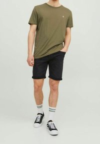 Hombre vistiendo una camiseta verde oliva, pantalones cortos de mezclilla negros arremangados, calcetines blancos con rayas grises y zapatillas negras y blancas, de pie de forma casual.