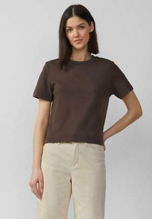 Braunes T-Shirt mit kurzen Ärmeln und rundem Halsausschnitt, aus weicher Baumwolle gefertigt. Gerader Schnitt mit minimalen Nähdetails, kombiniert mit hellcremefarbenen Hosen.