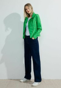 Veste en simili cuir vert avec un col à revers, associée à un jean en denim foncé à jambes larges et des baskets blanches. Design minimaliste et texture lisse.