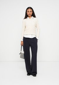 Crème kabelgebreide trui over een witte blouse, gecombineerd met marineblauwe wijde broek en een kleine handtas met patroon.