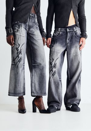 Dwie osoby ubrane w czarne prążkowane krótkie swetry oraz szare jeansy o szerokich nogawkach z czarnymi abstrakcyjnymi wzorami, zestawione z czarnymi butami.