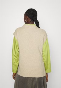 Frau mit geflochtenem Haar, die goldene Creolen trägt, einen beigen ärmellosen Pullover über einem limegrünen Langarmshirt und einen gefalteten metallischen Rock.