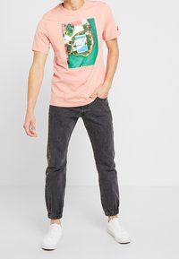 Camiseta de algodón rosa claro con un gráfico aéreo colorido de una cancha de baloncesto y la vegetación circundante. Combinada con pantalones oscuros con puños.