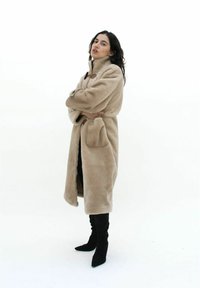 Beige fuskpälsjacka med hög krage, toggelstängningar och sidofickor, tillsammans med svarta överknäboots. Slät textur, knä-lång design.