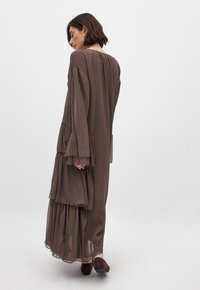 Abito lungo, marrone, a strati, realizzato in materiale leggero, con maniche ampie e vita arricciata. Indossato con scarpe scure e semplici.