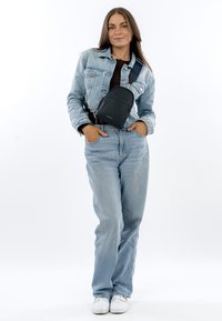 Junge Frau steht mit den Händen in den Taschen, trägt eine helle Jeansjacke und Jeans, eine schwarze Umhängetasche und weiße Turnschuhe vor weißem Hintergrund.