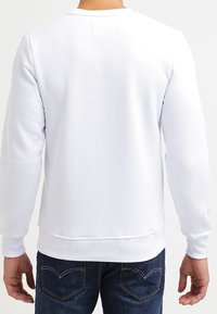 Weißer Pullover aus weichem Stoff, mit Rundhalsausschnitt und gerippten Bündchen. Getragen mit dunkelblauen Jeans, die ein einfaches, klares Design zeigen.