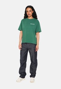 Camiseta de algodón verde con texto blanco "MULTIPLY", combinada con pantalones de mezclilla azul oscuro y zapatillas blancas. Cuello redondo, corte relajado.