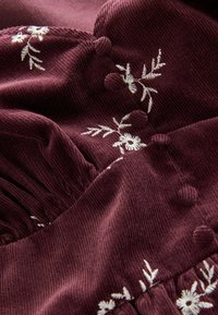 Le tissu de velours côtelé bordeaux présente de la broderie florale blanche et des boutons texturés, avec des éléments de design froncés créant profondeur et détail.