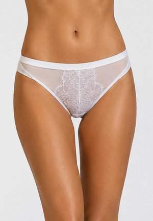 Kobieta nosząca prześwitujące białe koronkowe majtki bikini z kwiatowym haftem z przodu, odsłaniające środek ciała i górną część ud.