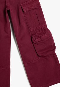 Pantaloni cargo color bordeaux con tasca a toppa e dettaglio di tessuto arrotolato sul lato, realizzati in un tessuto resistente con una consistenza liscia.