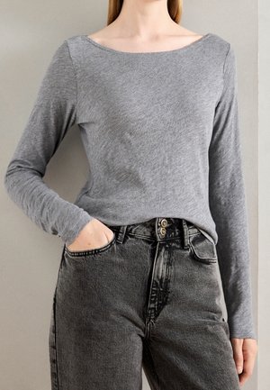 Kobieta ubrana w dopasowany szary top z długim rękawem włożony do wysokich, czarnych, wypranych dżinsów, z jedną ręką w kieszeni na jednolitym tle.