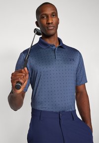 Golfové polo tričko v námořnické modři s jemným vzorem hvězd, s krátkými rukávy a límečkem. Spárováno s námořnickými kalhotami, drží golfovou hole.
