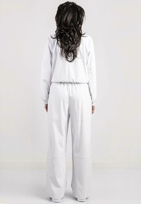 Salopette bianca a maniche lunghe con vita elastica e pantaloni a gamba larga, caratterizzata da una texture liscia e un design minimalista senza motivi o decorazioni.