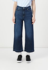 Blugi cu talie înaltă și croială largă, din denim închis, cu un design clasic cu cinci buzunare și detalii cu margine brută, asortați cu adidași negri.