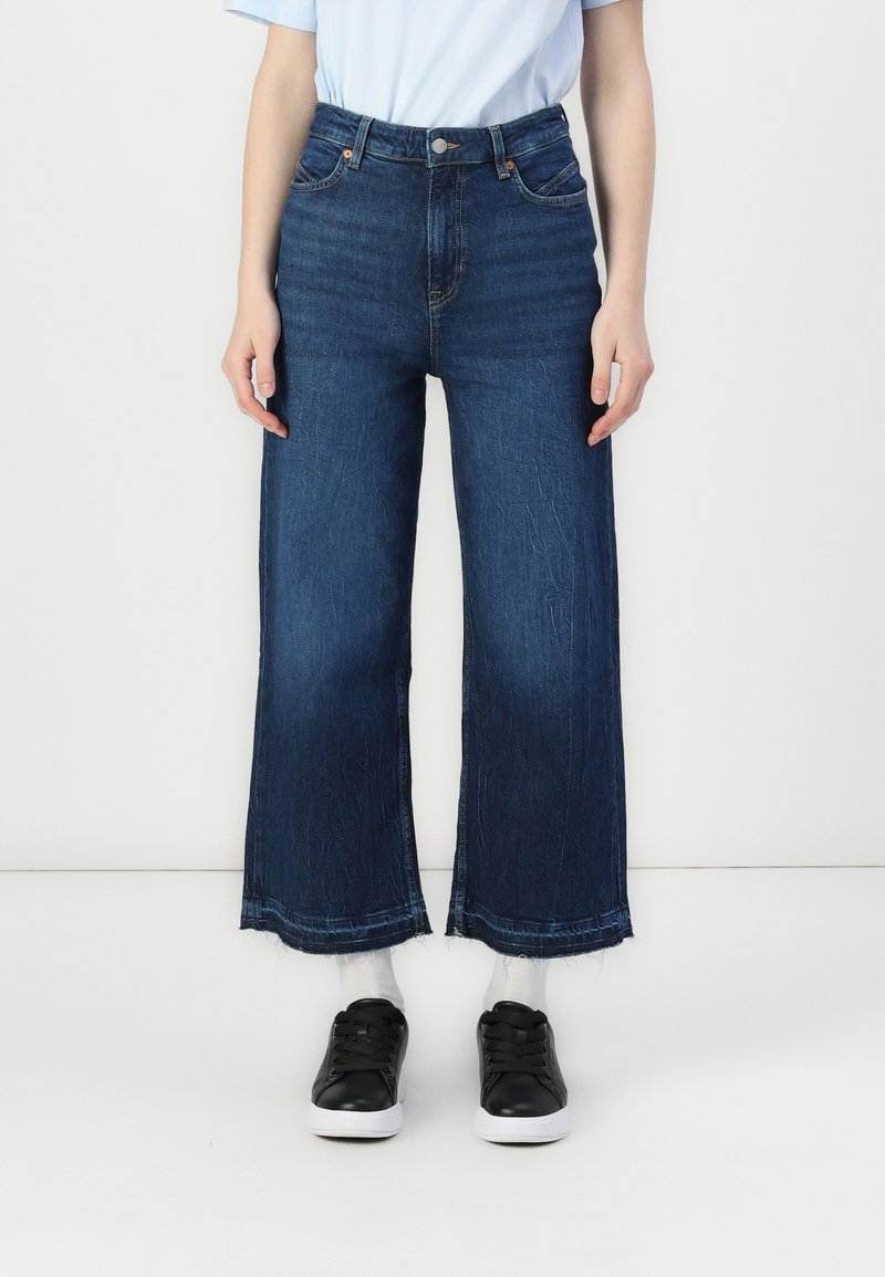Blugi cu talie înaltă și croială largă, din denim închis, cu un design clasic cu cinci buzunare și detalii cu margine brută, asortați cu adidași negri.