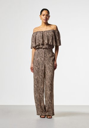 Vrouw staand in een off-shoulder zebra print jumpsuit en bruine open teen sandalen tegen een effen lichte achtergrond.