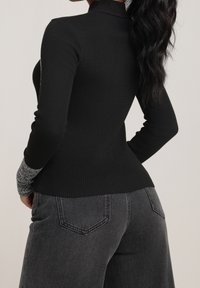 Personne portant un haut à manches longues noir côtelé avec des poignets gris et un jean noir délavé, vue de dos sur un fond uni.