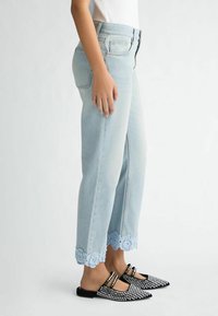Jeans taille haute bleu clair avec un détail de bord en dentelle scallopée. Associés à des ballerines noires à bout pointu avec des clous décoratifs.