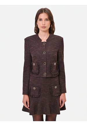 Donna che indossa un completo marrone scuro a due pezzi con giacca abbottonata e gonna abbinata con dettagli sulle tasche, in piedi su uno sfondo bianco.